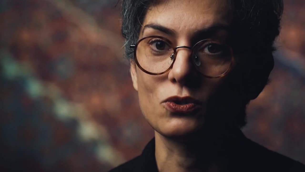 The woman, framed in a close-up, is speaking, her lips forming a slight pout. Her glasses reflect the light, and the background is a blur of warm colours.
