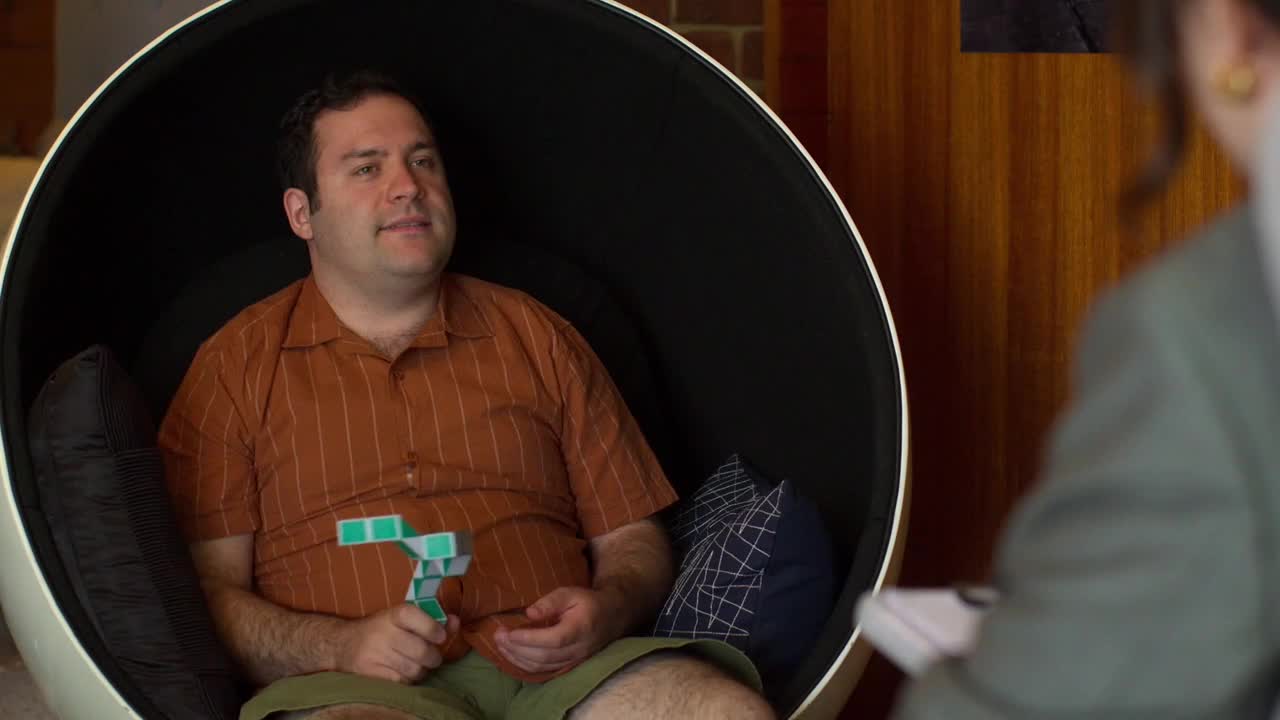 A man in an orange shirt sits in a black and white egg chair, holding a small, folded toy. He's looking to his right, toward someone out of frame.
