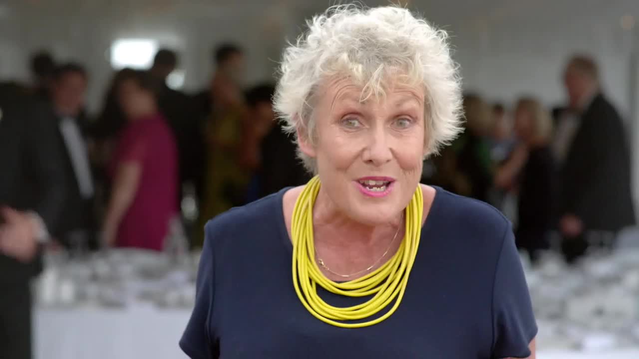 The woman in the foreground, wearing a bright yellow necklace, is speaking. Behind her, a crowd of people mill about, mostly out of focus.
