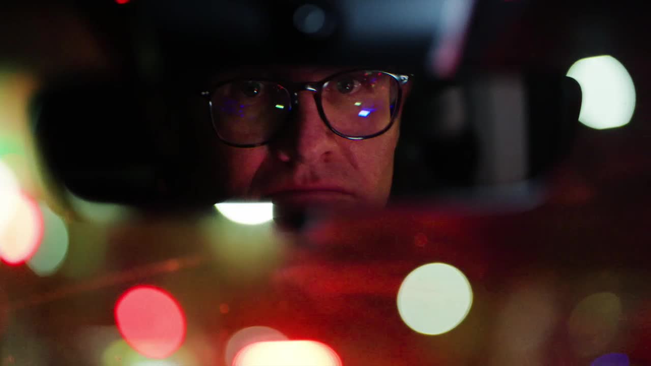 A man in glasses stares intently from the rearview mirror, his face illuminated by the red and white lights of the road. The blurred lights of other vehicles are reflected in his lenses.
