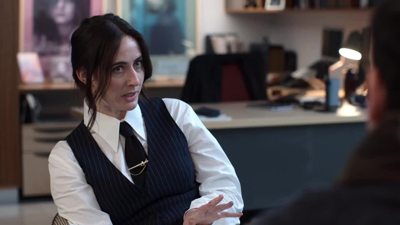 A woman in a pinstripe vest gestures with her right hand as she speaks. The other person is mostly out of view, but the woman seems to be engaged in a serious conversation.
