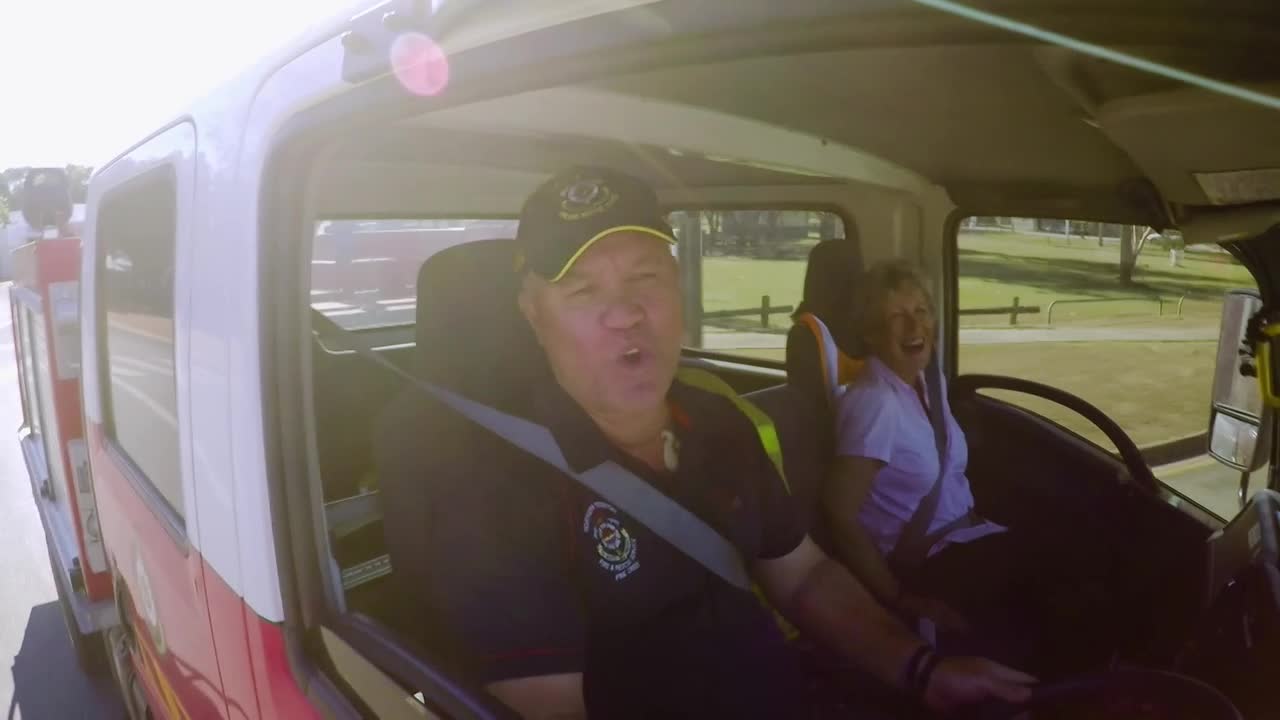 The man in the driver's seat is speaking, his mouth open, as the vehicle moves along a road. A woman in the passenger seat is laughing, and the sun shines through the windows of the vehicle.
