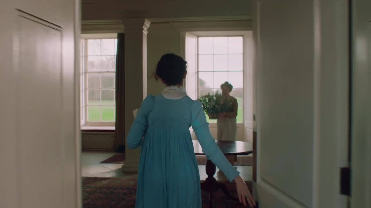 A woman in a light blue dress moves through a doorway, her hand reaching out to steady herself. In the distance, another woman stands by a table, holding a large basket of greenery near a window.
