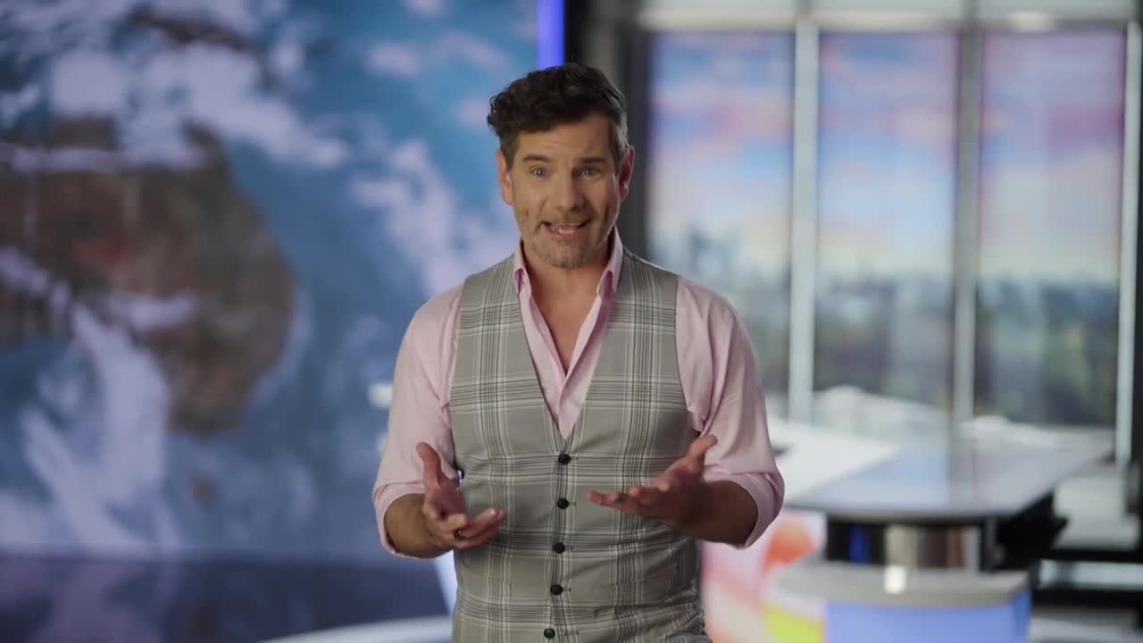 The man in the waistcoat gestures expressively, speaking to the camera. Behind him, a map of Australia and the world forms the backdrop of a news studio.
