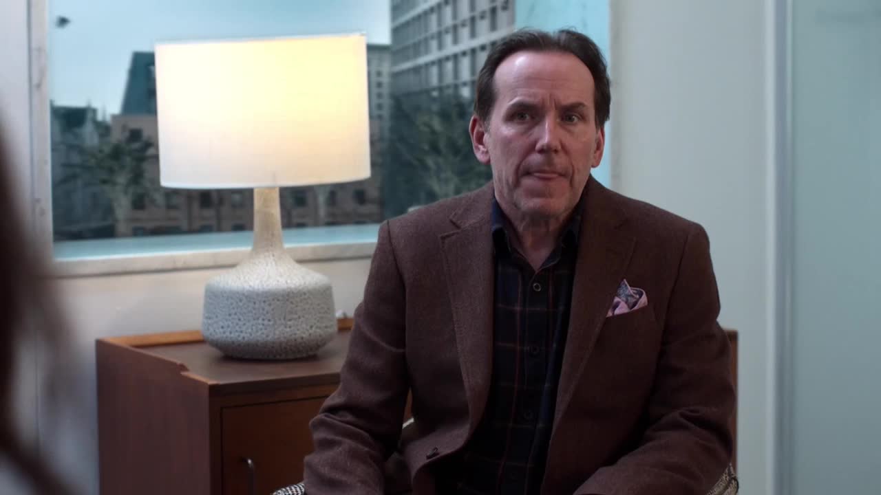 A man in a brown jacket and plaid shirt sits in front of a window. The lamp on the side table casts a warm glow on the room.
