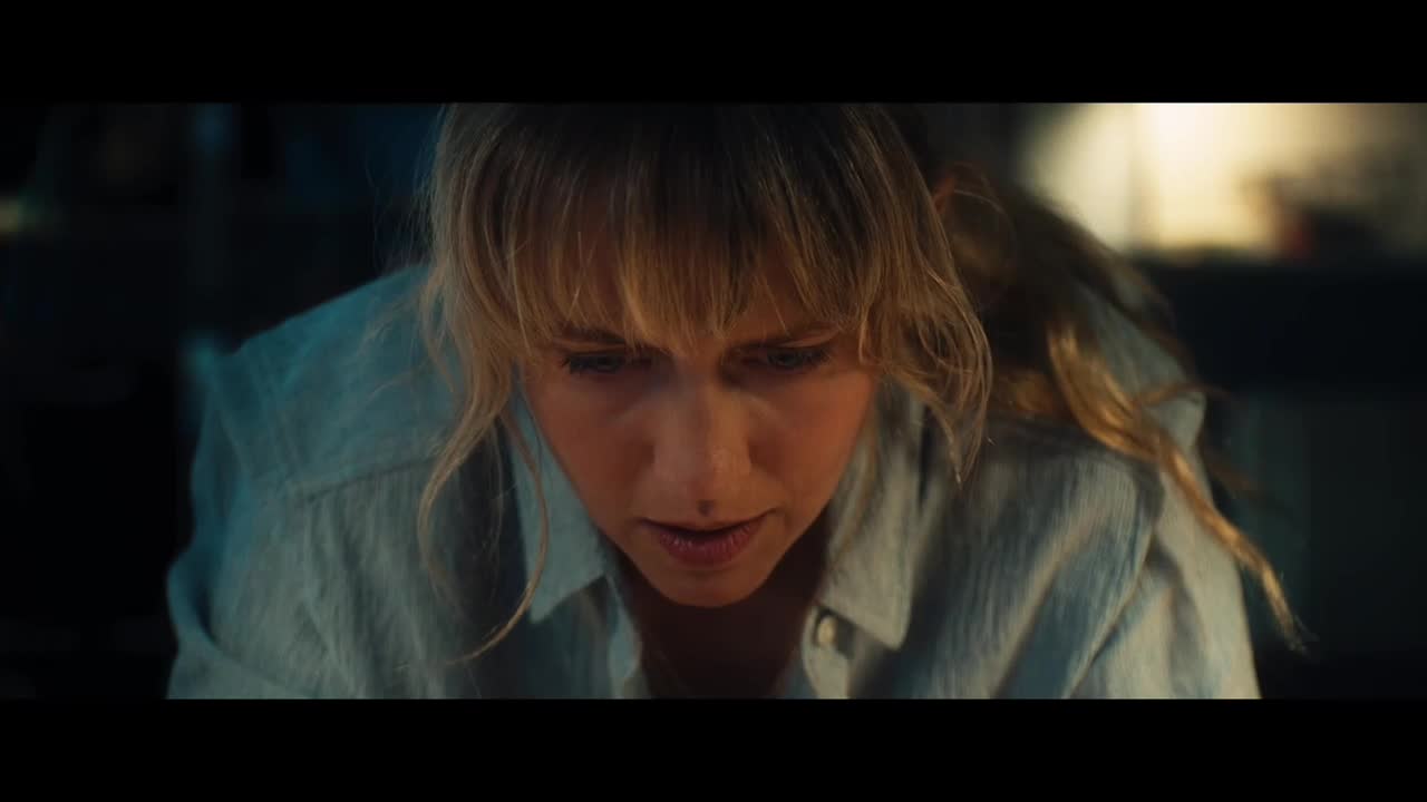 A woman with blonde hair and a light blue shirt is looking down with a concerned expression. The lighting is dim, and her face is illuminated.
