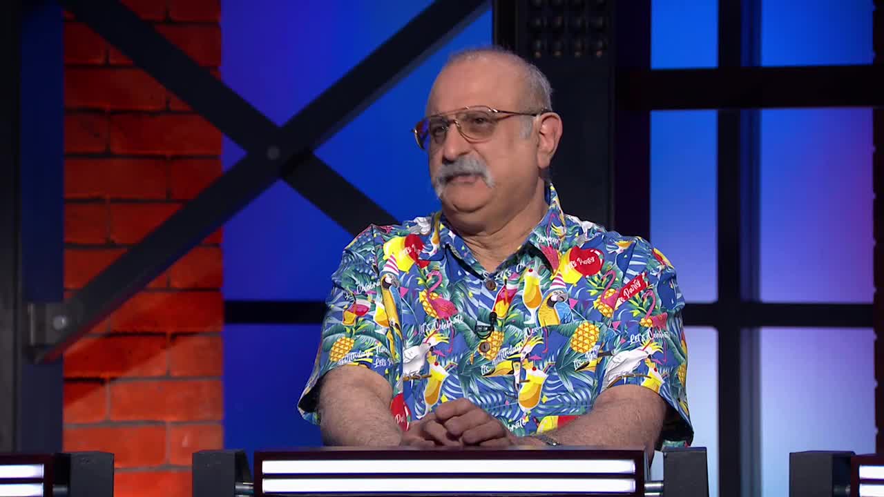 A man in a colorful shirt and glasses sits at a table, looking towards the camera. The background is a mix of brick and blue, with a metal crossbar.
