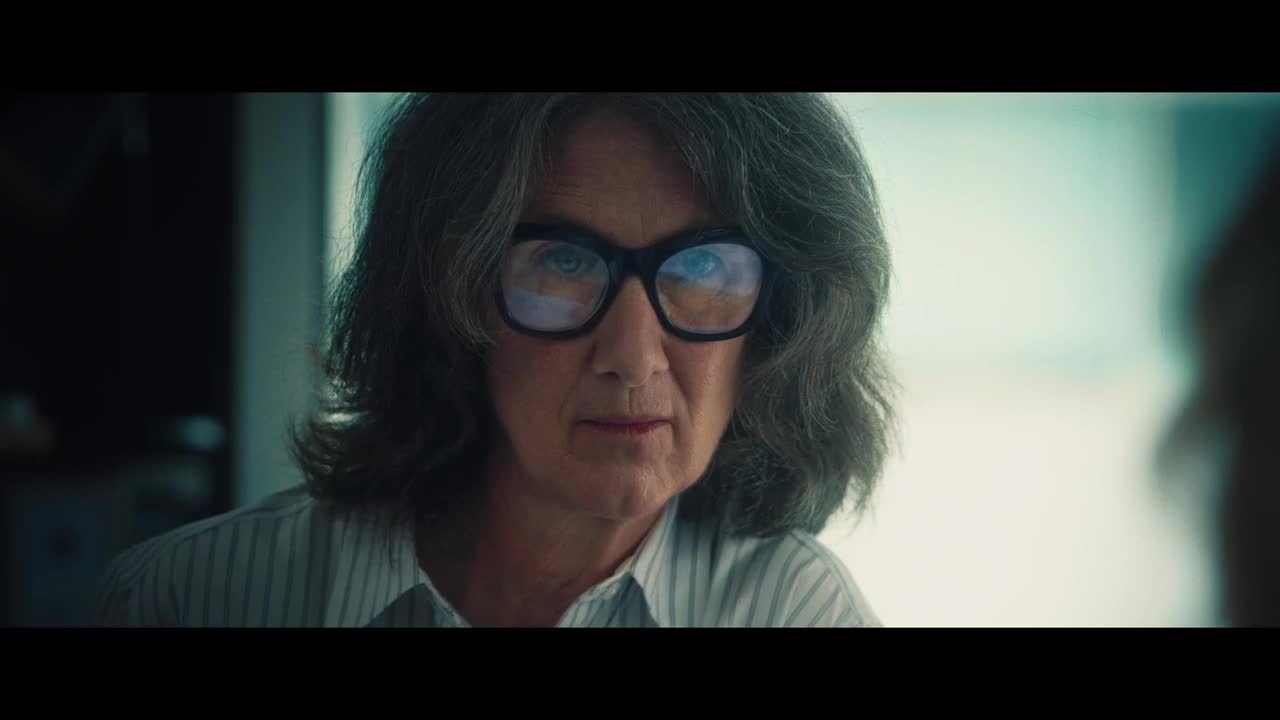 A woman with graying hair and thick-rimmed glasses stares directly ahead, her expression unreadable. The light reflects off her lenses, and the pale stripes of her shirt are visible.
