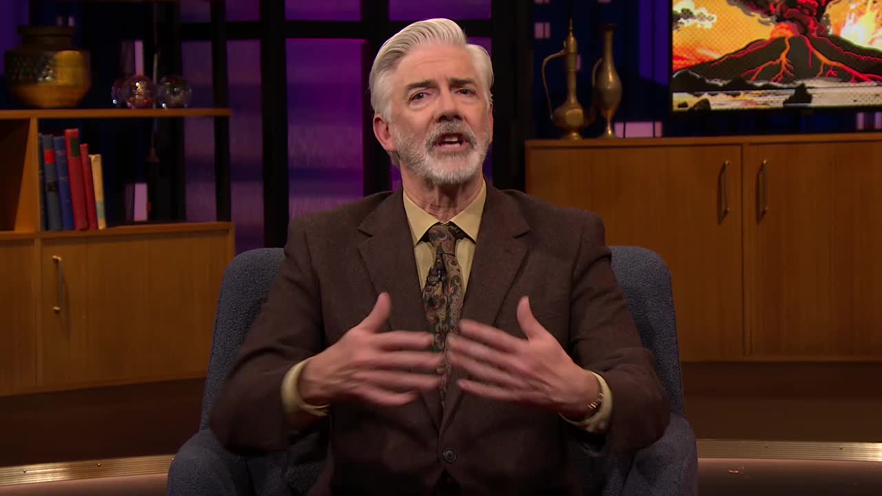 The man in the brown suit gestures with both hands, his expression animated. Behind him, a colorful backdrop and shelves filled with objects create a studio setting.
