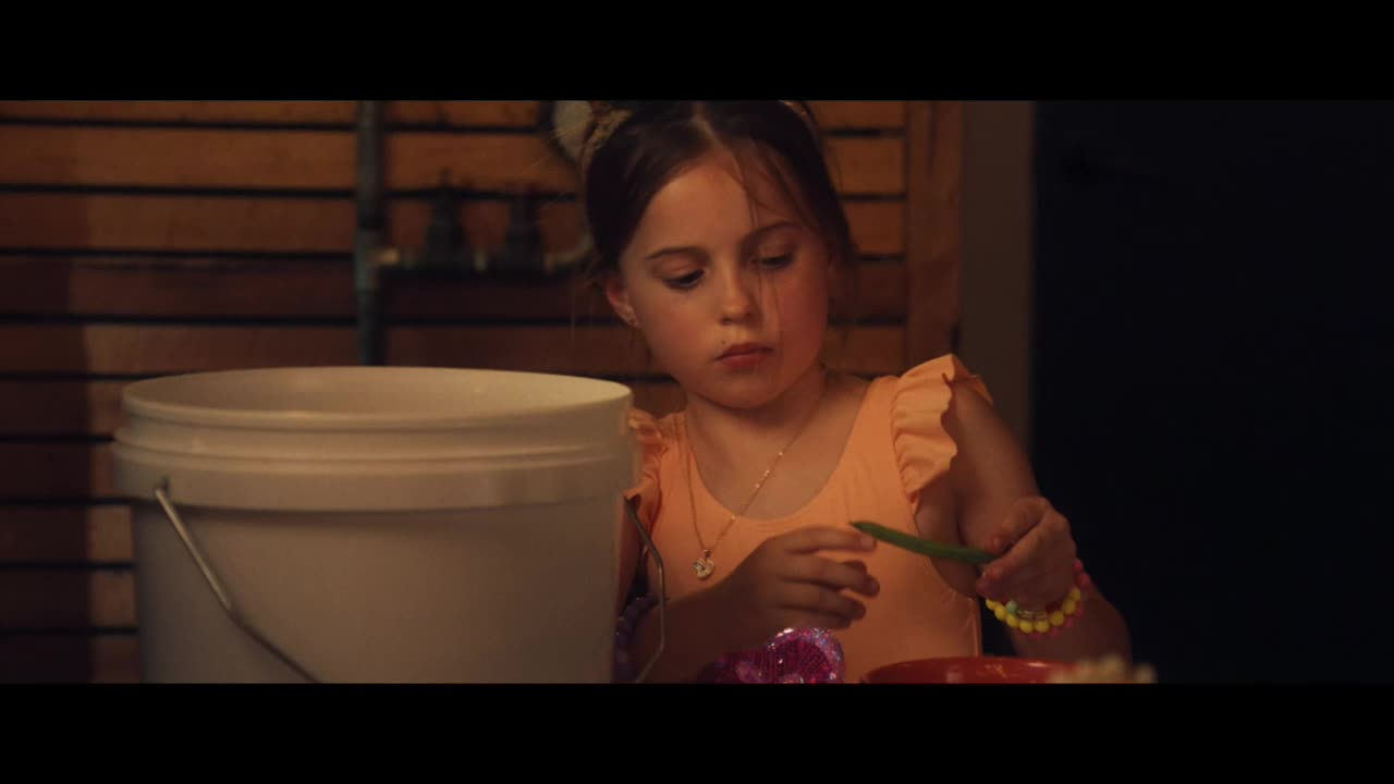 A young girl, wearing a peach-colored top, carefully holds a green bean. She's sitting at a table, with a white bucket and a plate of colorful items nearby.
