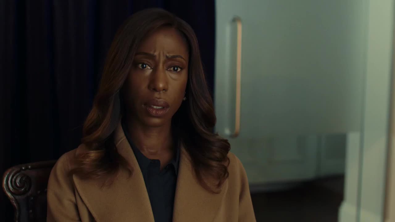 Her brow is furrowed, and her lips are slightly parted as she looks directly ahead. The woman is wearing a tan coat over a dark blue shirt, and her gaze seems troubled.
