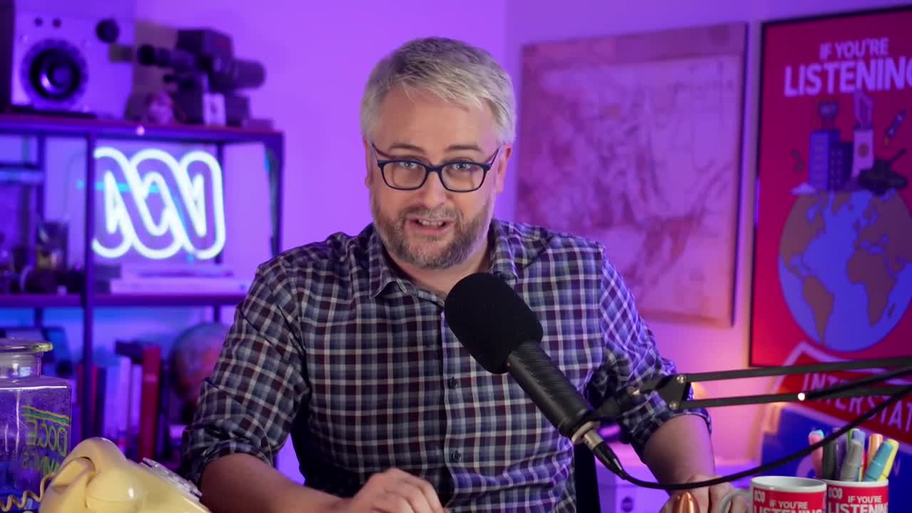 He's speaking directly into a microphone, his checkered shirt and glasses catching the light. Behind him, the ABC logo glows, and a poster reads, "If You're Listening."
