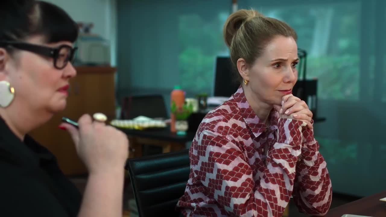 The woman in the patterned shirt has her hands clasped under her chin, looking concerned. Beside her, the other woman holds a pen, her expression equally serious.
