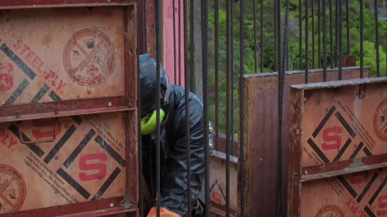 A worker, clad in a dark rain jacket and neon green ear protection, is hunched over, working within a framework of steel rebar and wooden forms. The construction site is surrounded by the lush green of the Australian bush.
