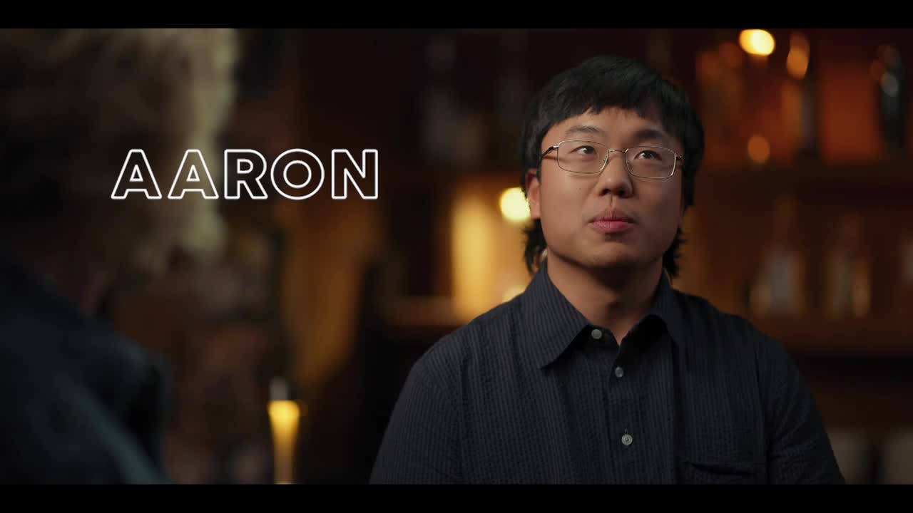 "Aaron" is spelled out in a glowing font. A man in glasses and a dark shirt looks toward someone off-screen, his expression thoughtful.
