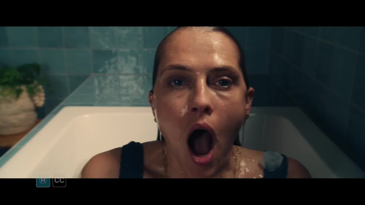 A woman sits in a bathtub, her face contorted in a wide, silent scream. Water streams down her face, and she appears to be in distress.
