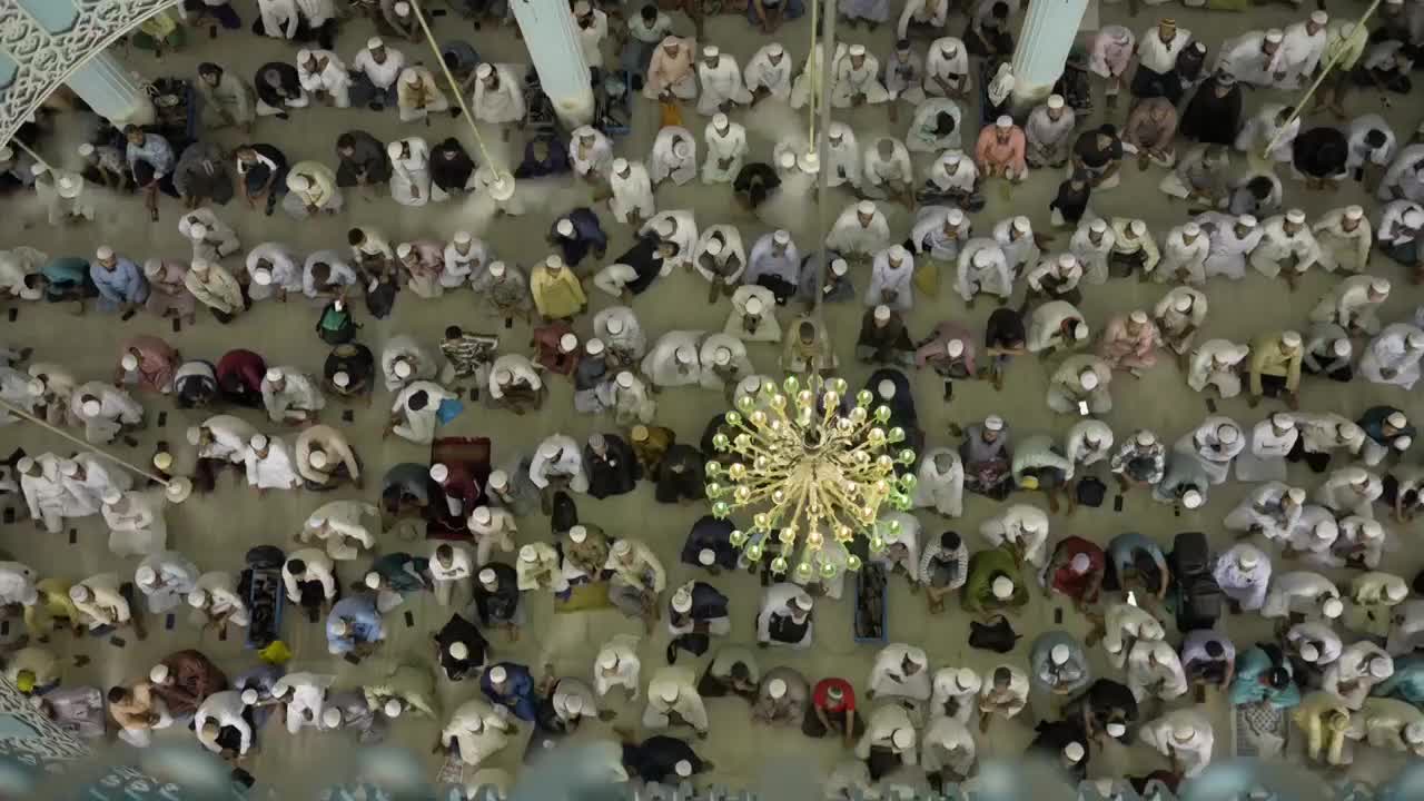 Hundreds of men, dressed in white and various colours, are bowing in unison on the floor. A large chandelier hangs above them, illuminating the scene.
