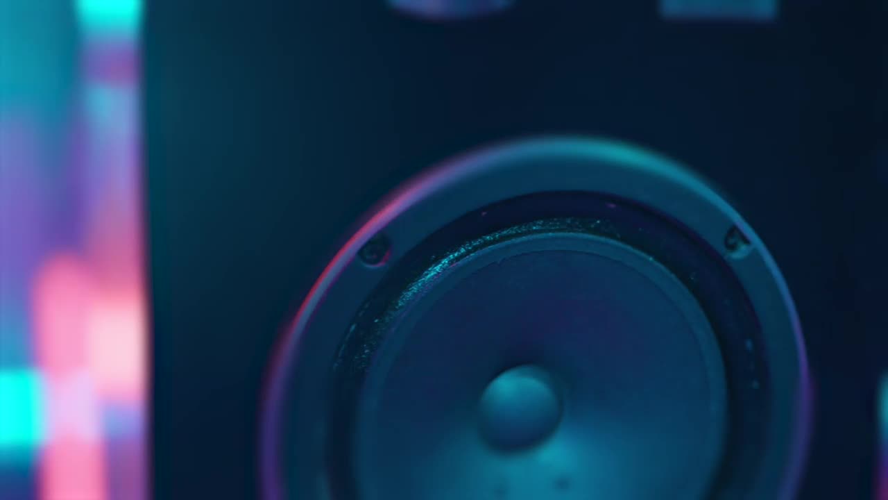 A speaker cone sits in the foreground, its dark surface reflecting the blue and purple hues of the surrounding lights. The small, circular holes around the rim of the speaker are visible.
