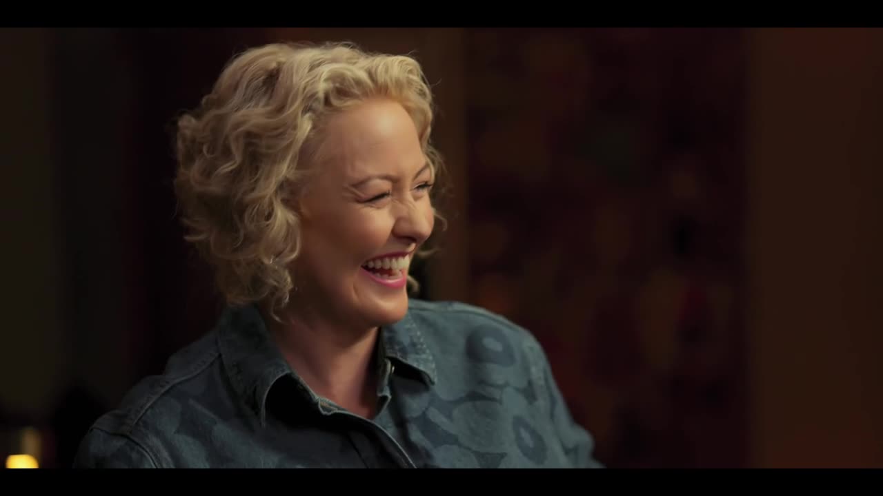 A woman with curly blonde hair is laughing, her head tilted back slightly. She's wearing a patterned denim shirt, and the light catches her face as she smiles.
