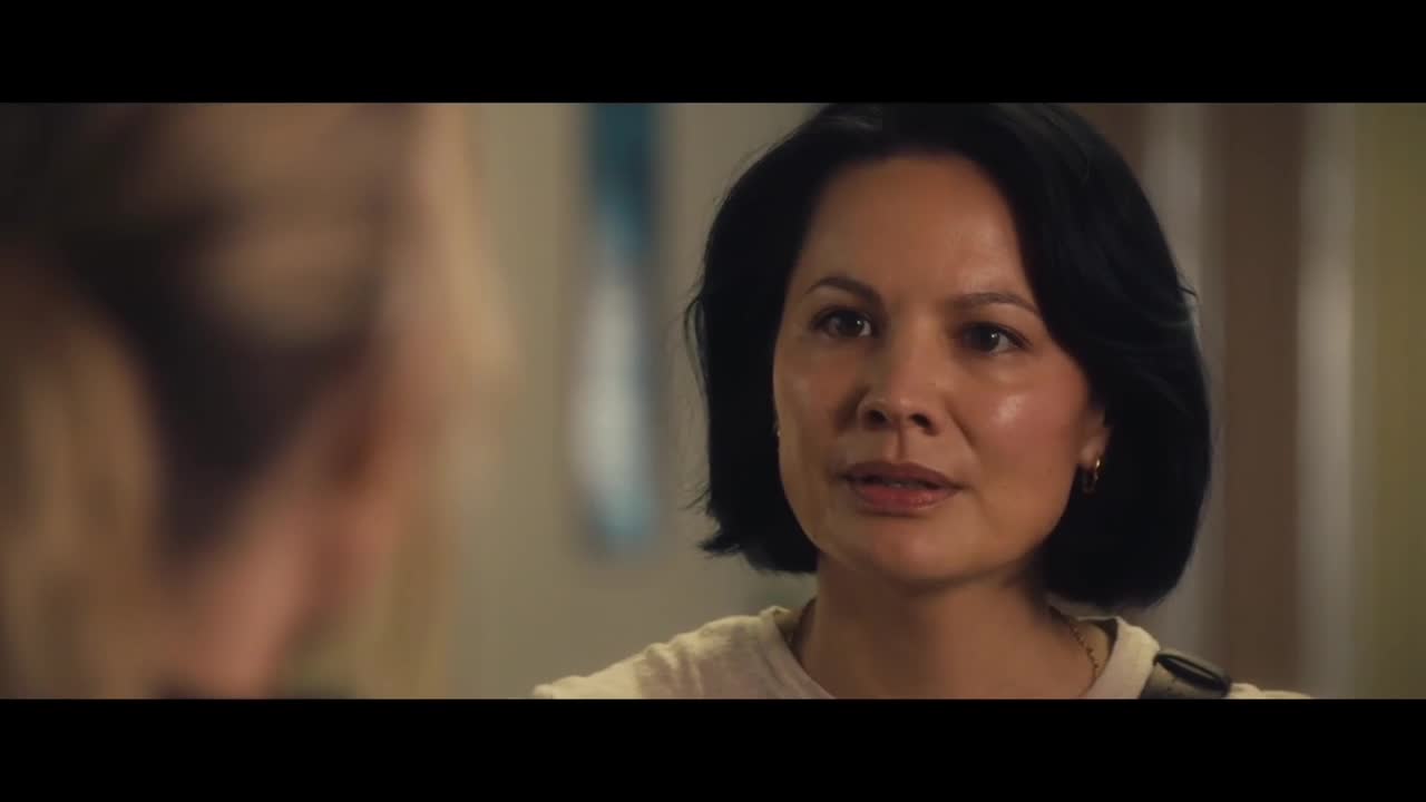 A woman with dark hair and a cream-colored top is speaking, her expression serious. The other person is out of focus, but the woman is clearly engaged in a conversation.
