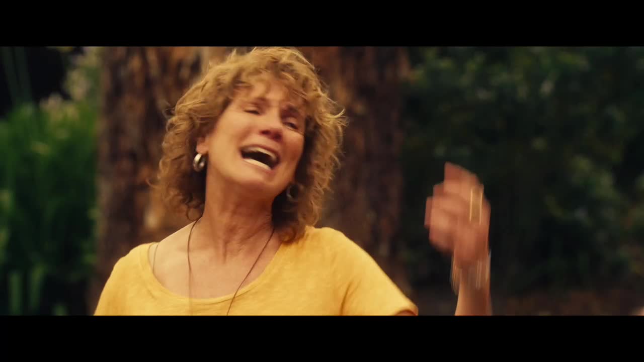 A woman in a yellow shirt throws her arms up, mouth open in what looks like a scream, as if she's just received some shocking news. Her curly hair bounces as she moves, and her expression is one of pure emotion.
