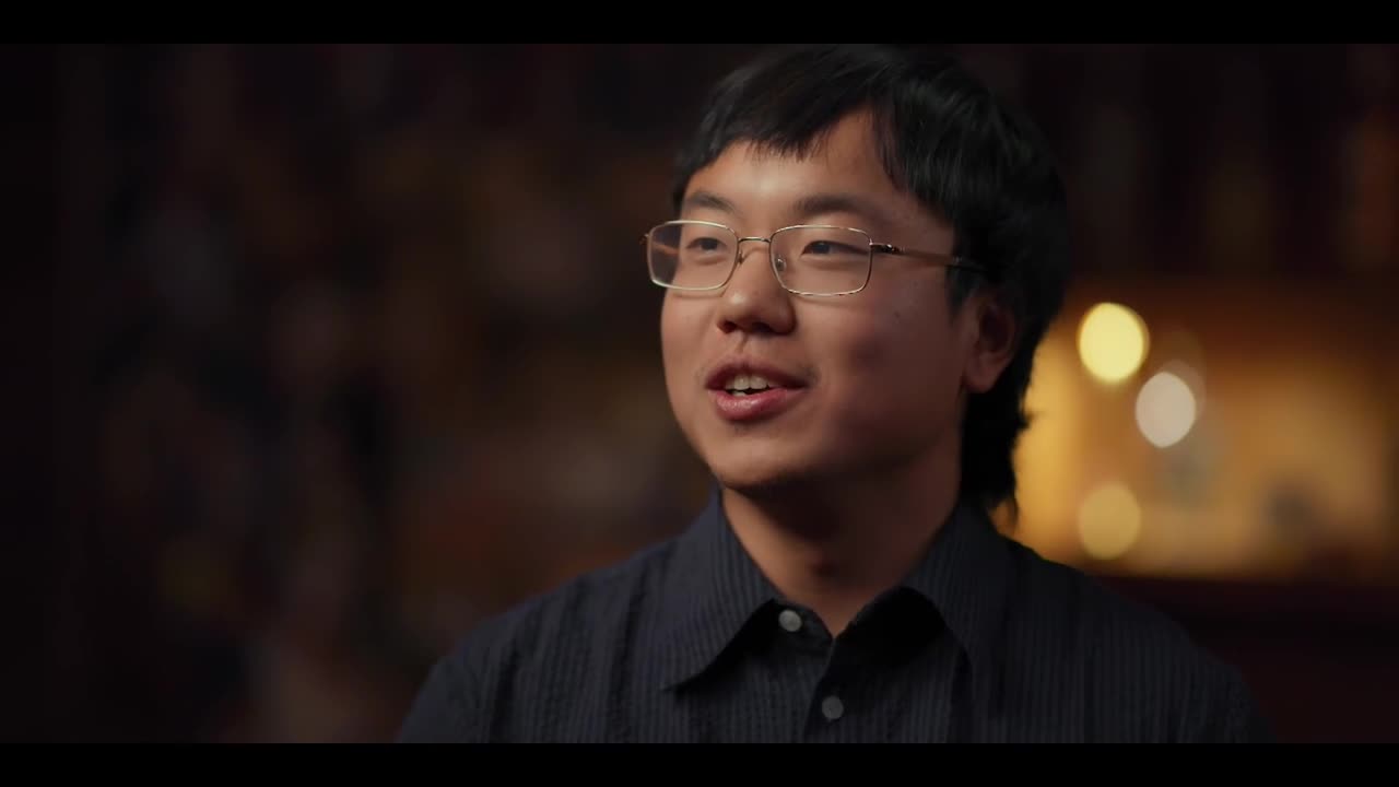 The man with glasses is speaking, his mouth moving as he talks. Soft light illuminates his face against a blurred backdrop.
