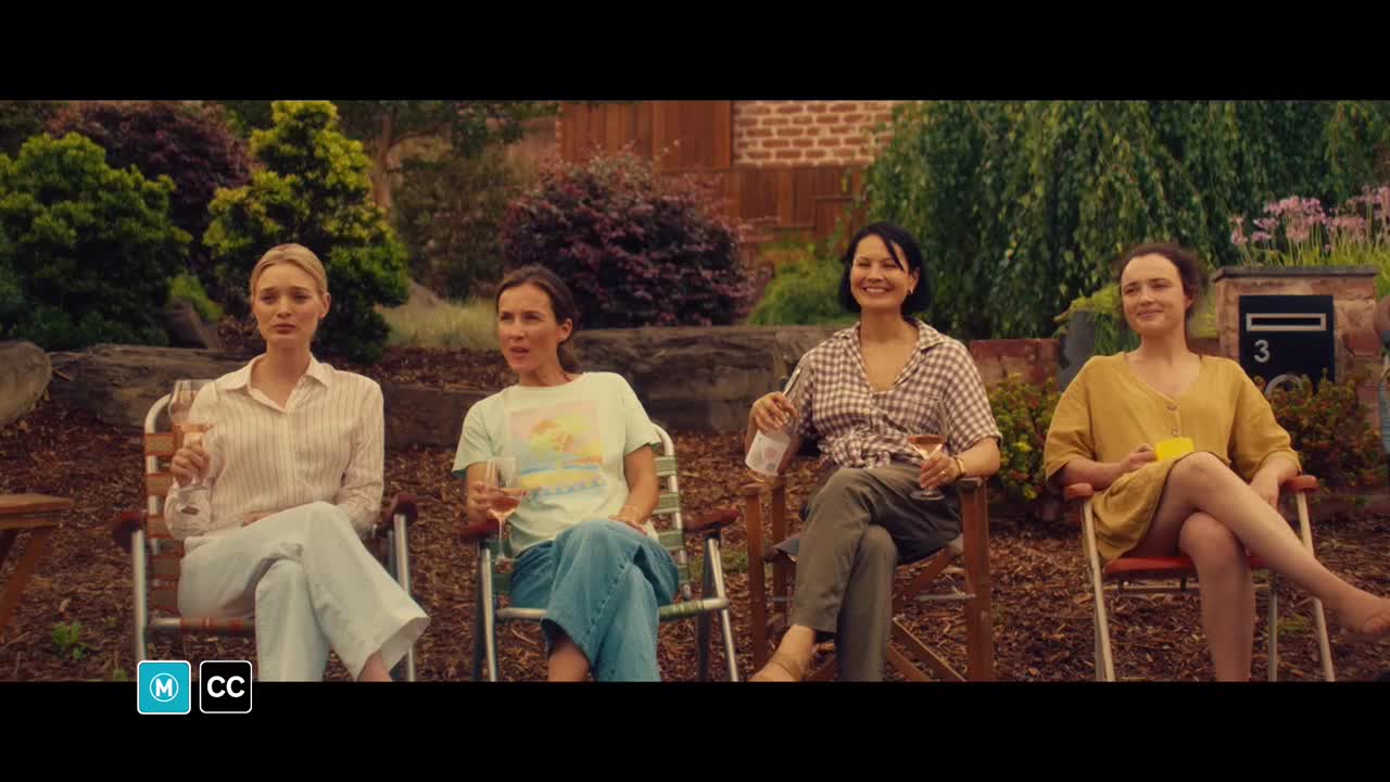 Four women sit in folding chairs, each holding a glass of wine, chatting in a backyard. The sunlight catches the pink in their drinks as they lean back, relaxed.
