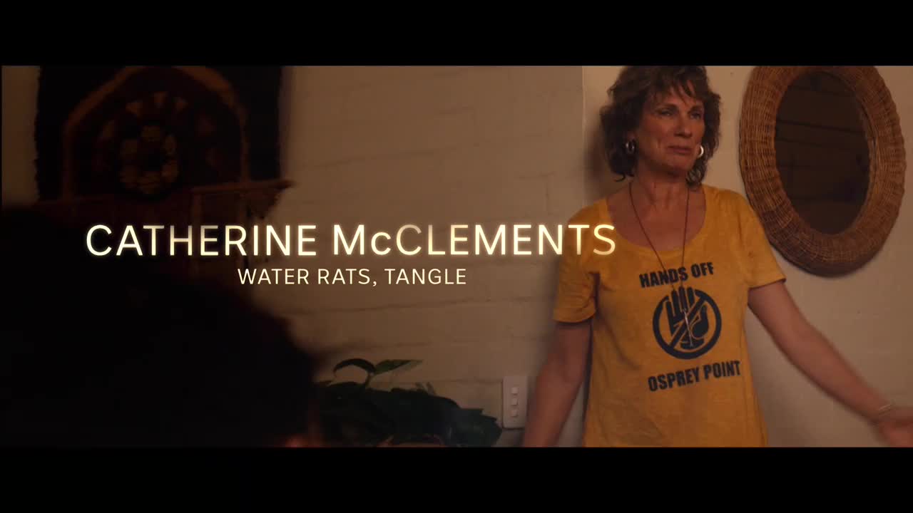 Catherine McClements, of *Water Rats* and *Tangle* fame, stands in a doorway, wearing a yellow t-shirt with a protest message. She gestures with her hands, as if about to speak.
