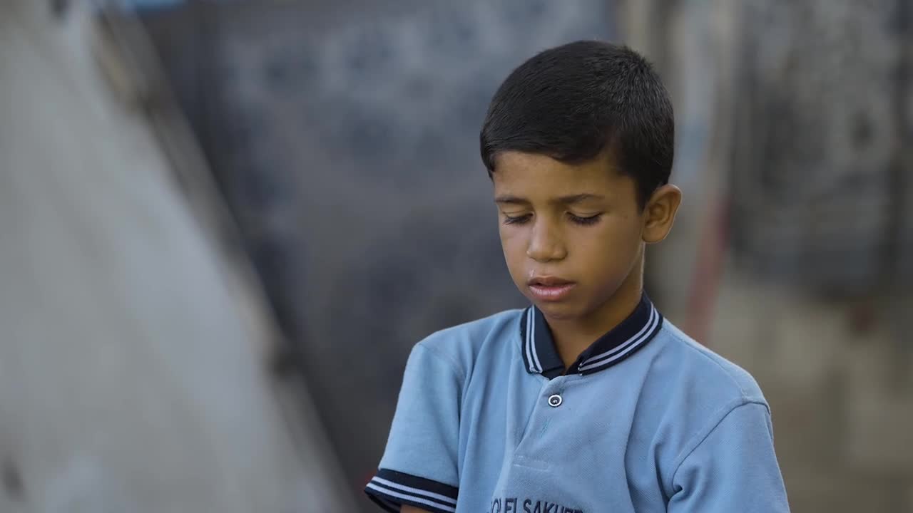 A young boy, wearing a blue school uniform, looks down with a troubled expression. His eyes are downcast, and a trickle of something is visible below his lip.
