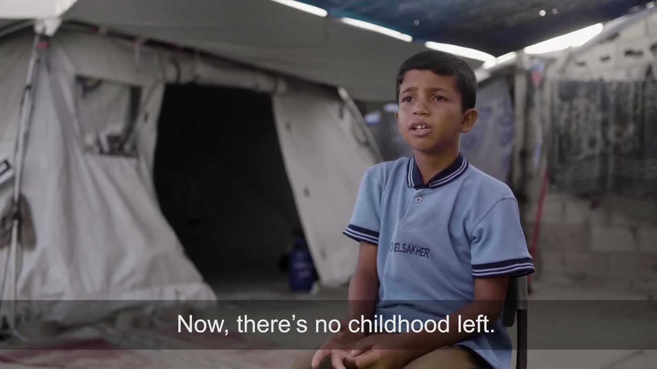 A young boy in a blue school shirt is speaking, his gaze directed slightly upward. Behind him, a tent and makeshift structures suggest a refugee camp, the backdrop to his words.
