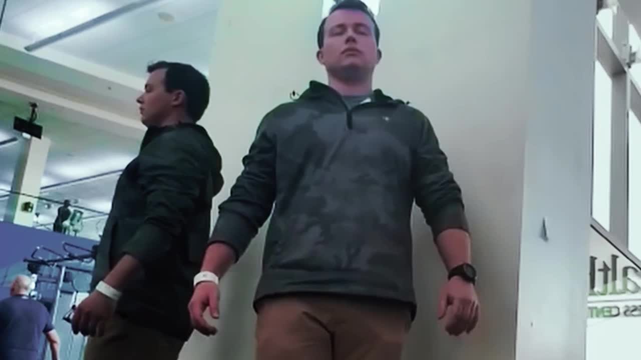 A man in a gray hoodie stands with his eyes closed, arms at his sides, leaning against a white wall. Beside him, a mirror image of the man stands in the same pose.
