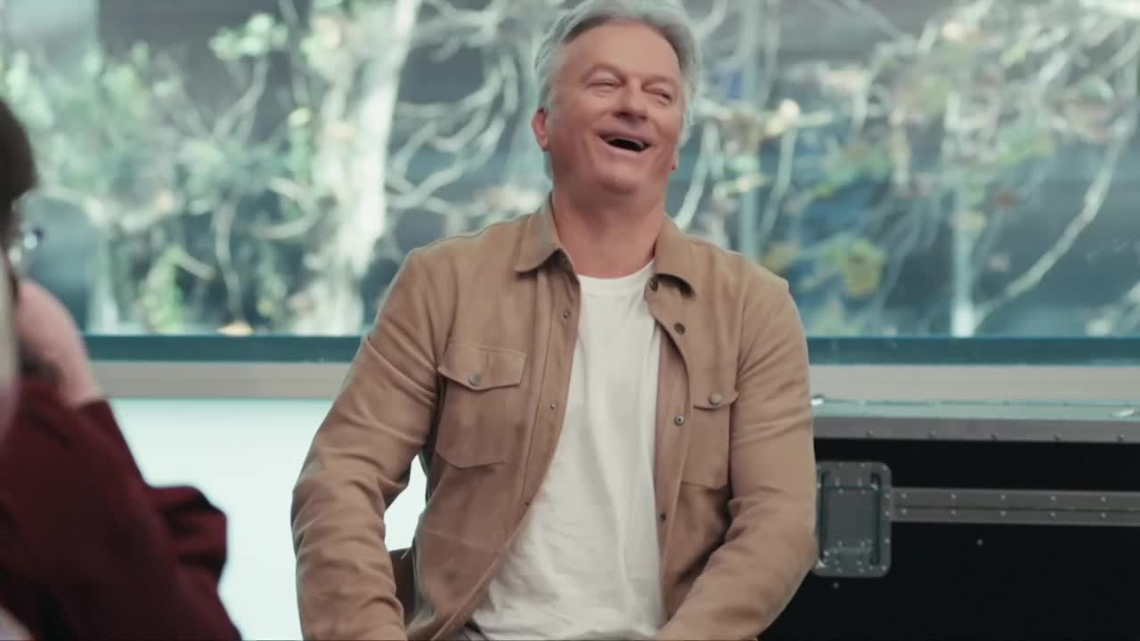 The man in the tan shirt is laughing, his head thrown back as he sits in front of a window. Beside him, someone in a burgundy jacket is partially visible.
