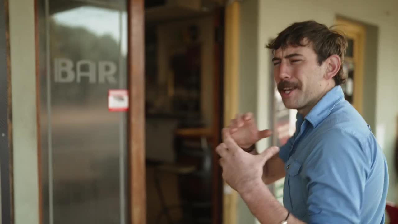 A man in a blue shirt gestures emphatically, his hands framing an invisible shape. He's standing outside a bar, the word "BAR" visible on the glass door behind him.

