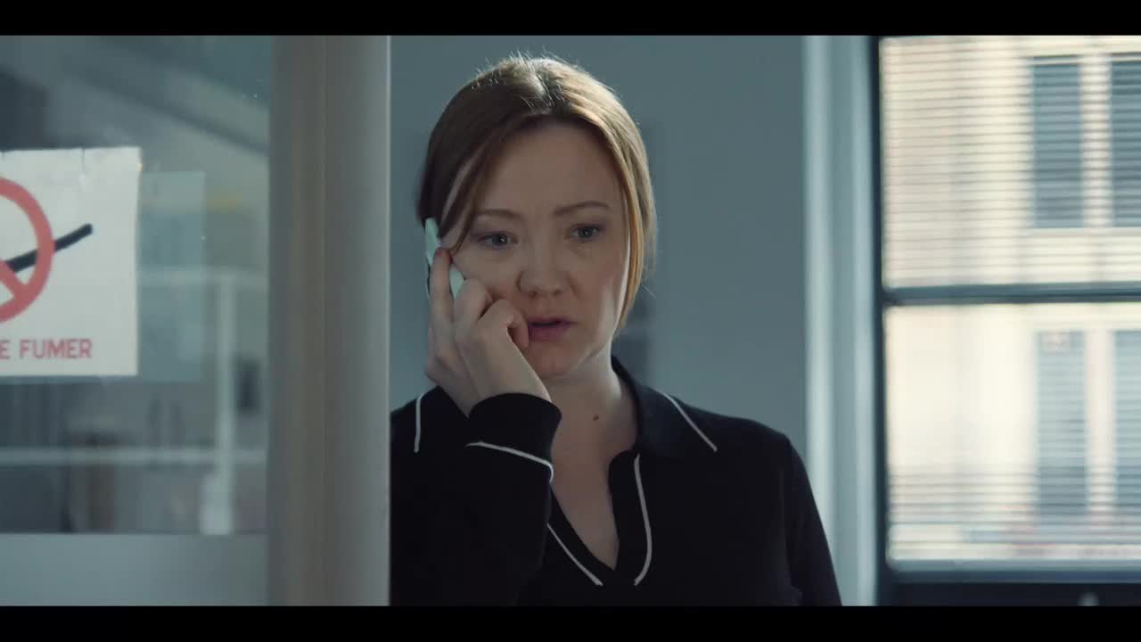 She's holding a phone to her ear, listening intently, her expression a mix of concern and concentration. The no smoking sign on the glass door behind her is a stark contrast to the modern office setting.
