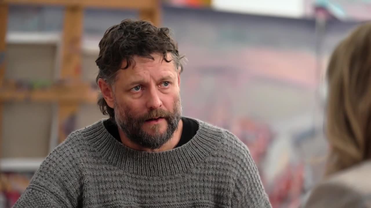 A man with a beard and curly hair, wearing a gray sweater, is looking intently at someone off-camera. Behind him, a colorful painting is visible, suggesting an interview on ABC Australia.
A man with a beard and curly hair, wearing a gray sweater, is looking intently at someone off-camera. Behind him, a colorful painting is visible, suggesting an interview on ABC Australia.