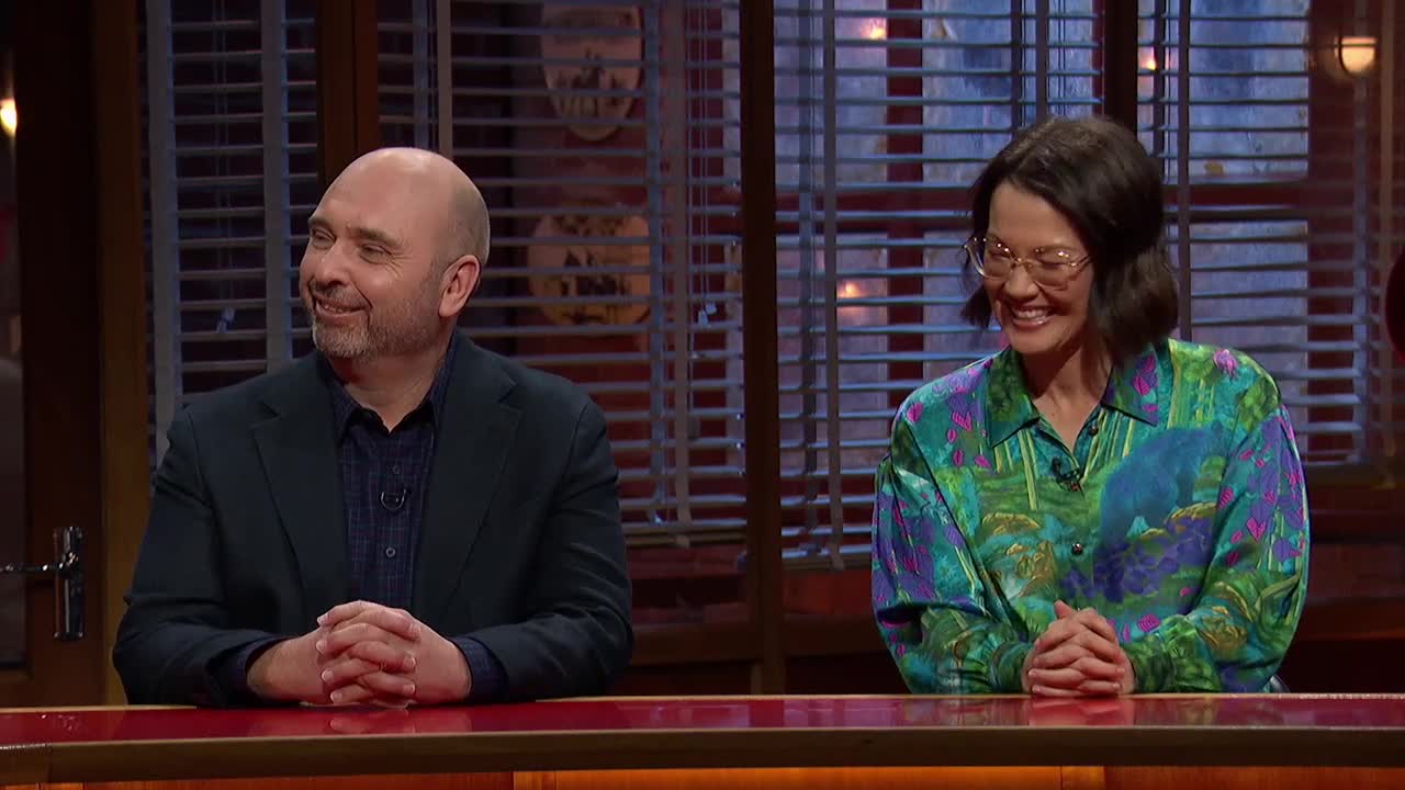 Two people sit behind a red desk, smiling. The woman in the patterned shirt and glasses looks toward the man, who is also smiling, and they both have their hands clasped.
Two people sit behind a red desk, smiling. The woman in the patterned shirt and glasses looks toward the man, who is also smiling, and they both have their hands clasped.