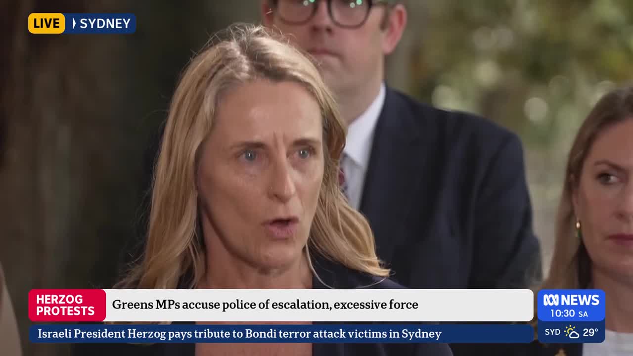 A woman with blonde hair speaks intently, her mouth open. Behind her, a man in a suit and glasses stands, his gaze directed forward.