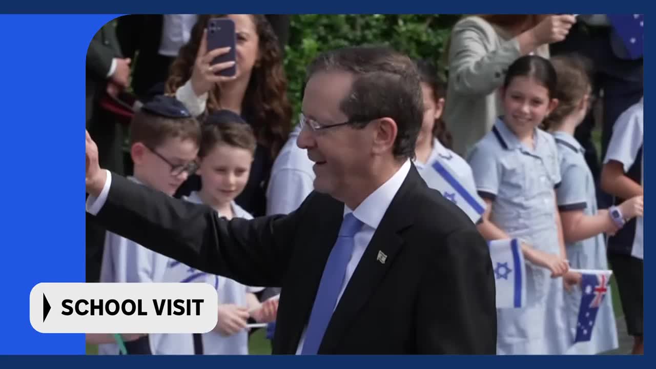 A man in a suit waves to a crowd of children holding small Israeli and Australian flags. The children, some wearing kippahs, are lined up, watching him approach.