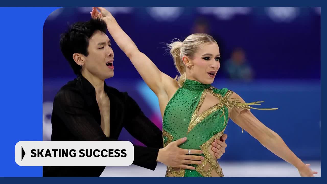A male figure skater in a black shirt holds his female partner, who wears a sparkling green costume. Their arms are raised as they glide across the ice.