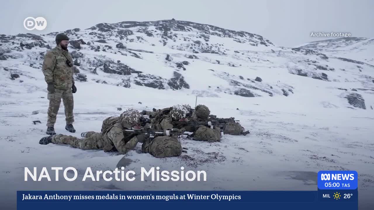 Soldiers in camouflage lie prone in the snow, aiming their rifles forward. Another soldier stands behind them, observing the snowy, rocky terrain.