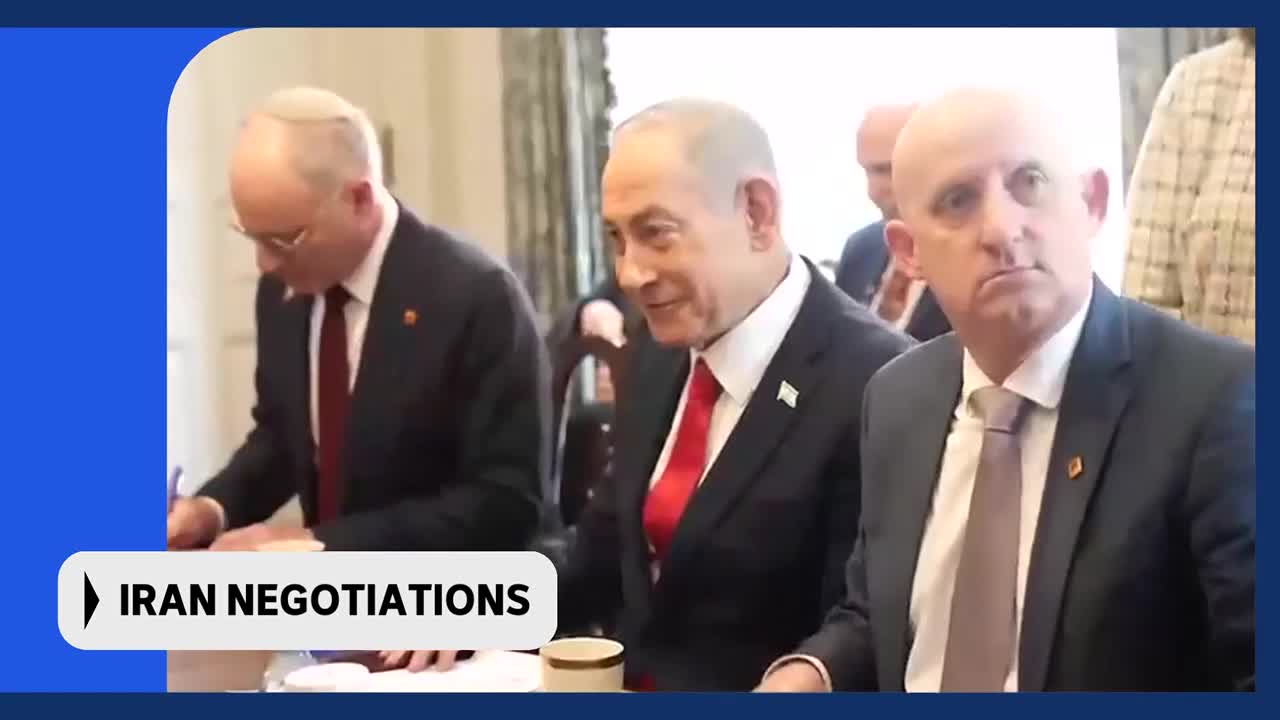 A man in a dark suit and red tie smiles as he looks to his left. Beside him, another man in a suit signs a document.