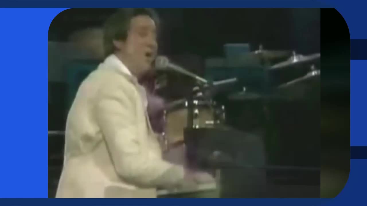 A man in a cream jacket sings into a microphone while playing a keyboard. Behind him, a drummer is partially visible, poised over his kit. A man in a cream jacket sings into a microphone while playing a keyboard. Behind him, a drummer is partially visible, poised over his kit.