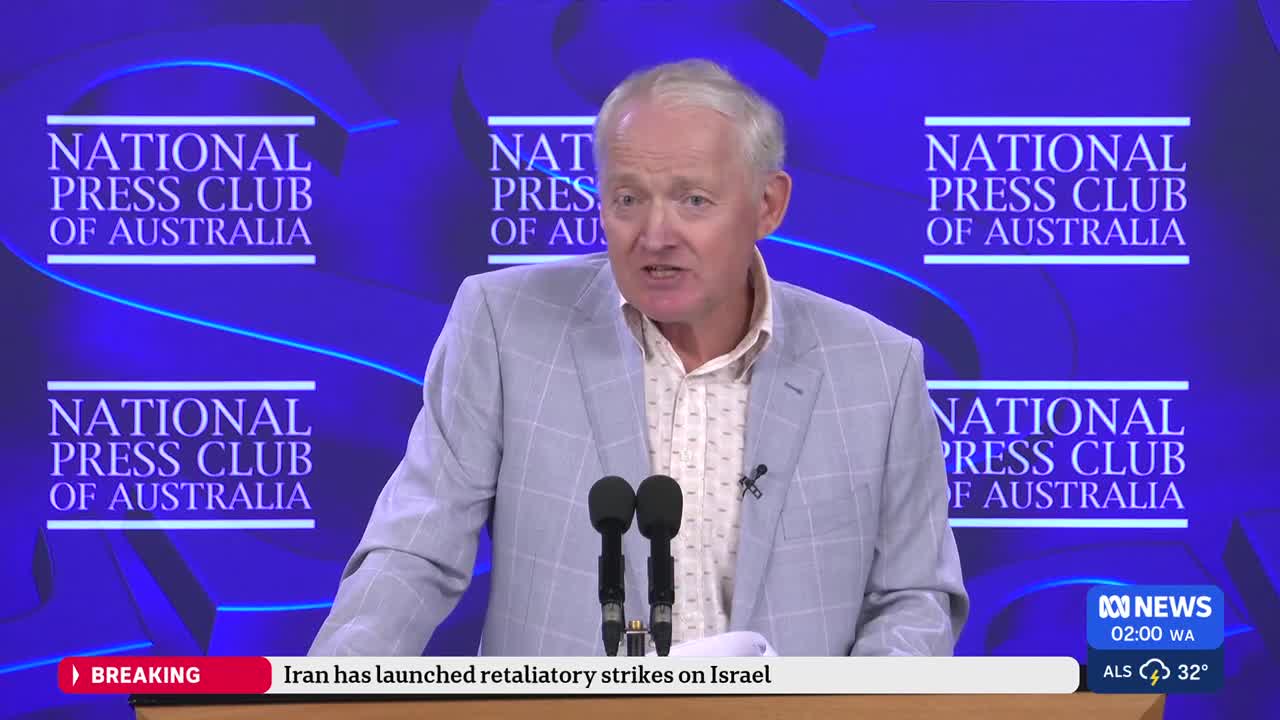 A man in a light grey suit stands at a podium, speaking into microphones. The backdrop behind him is a deep blue with repeating logos for the National Press Club of Australia. A news ticker at the bottom of the screen reads "Iran has launched retaliatory strikes on Israel." A man in a light grey suit stands at a podium, speaking into microphones. The backdrop behind him is a deep blue with repeating logos for the National Press Club of Australia. A news ticker at the bottom of the screen reads "Iran has launched retaliatory strikes on Israel."