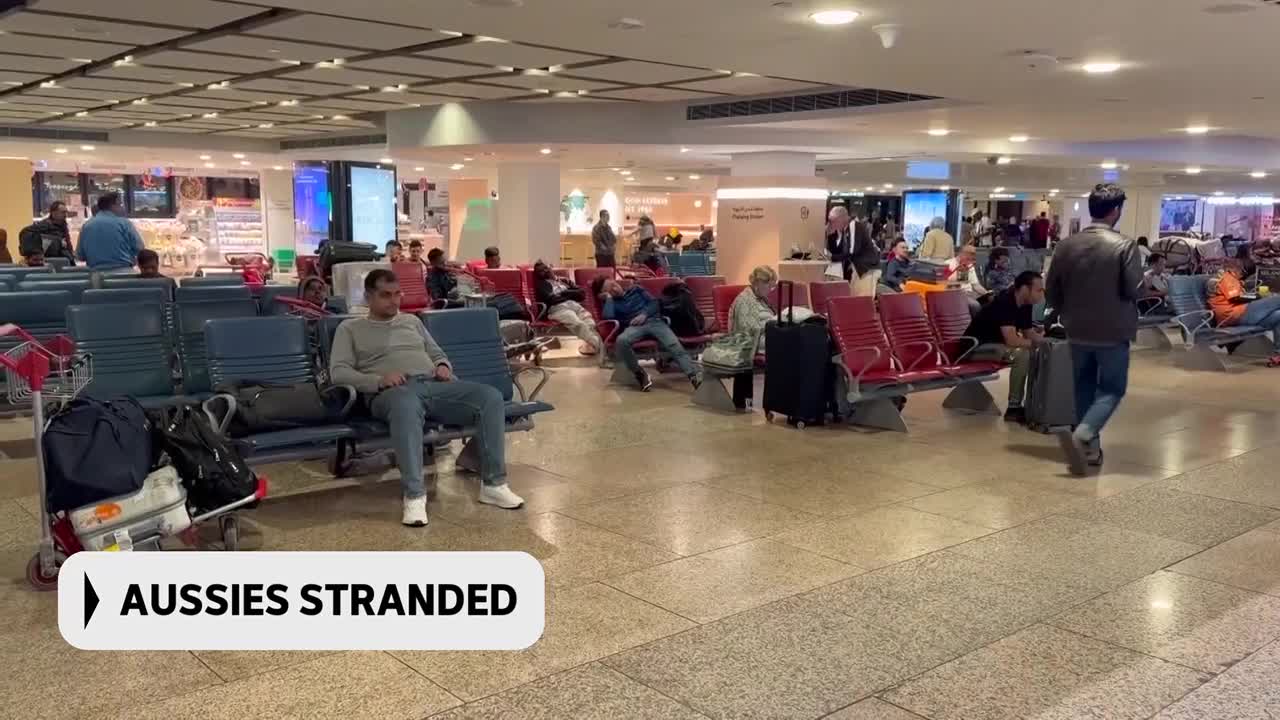 People are sitting in rows of airport chairs, some with luggage at their feet. A man walks past with a suitcase, heading towards the departure gates. People are sitting in rows of airport chairs, some with luggage at their feet. A man walks past with a suitcase, heading towards the departure gates.