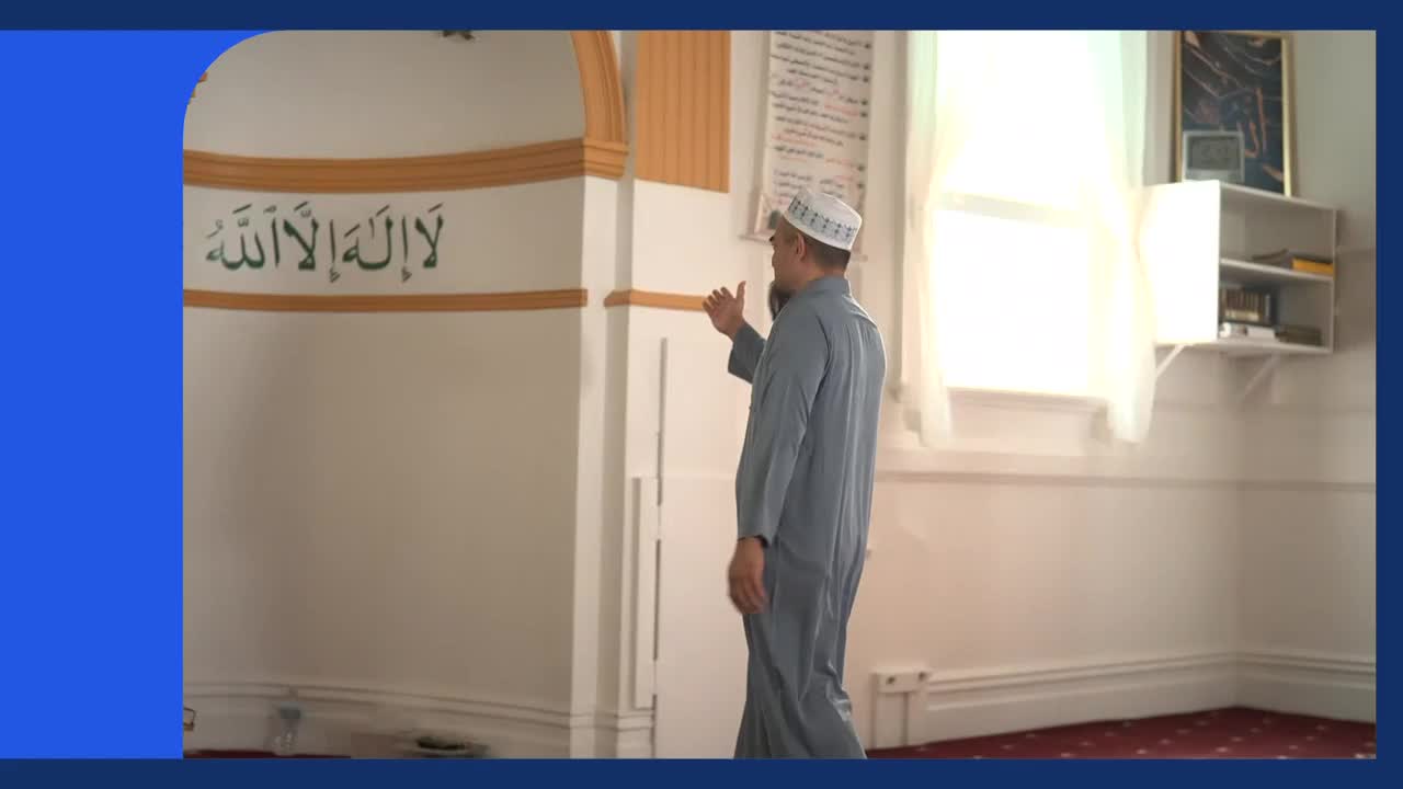 A man in a grey robe and white cap walks past Arabic calligraphy on a mosque wall. Sunlight streams through a window, illuminating the room. A man in a grey robe and white cap walks past Arabic calligraphy on a mosque wall. Sunlight streams through a window, illuminating the room.