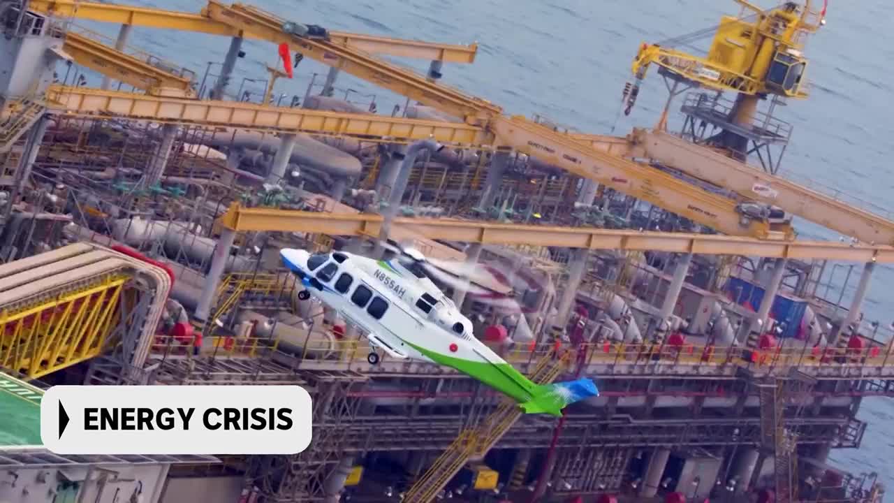 A white and green helicopter hovers near a sprawling offshore oil rig. The rig, a complex network of yellow cranes and metal structures, sits on the water under a cloudy sky. A white and green helicopter hovers near a sprawling offshore oil rig. The rig, a complex network of yellow cranes and metal structures, sits on the water under a cloudy sky.