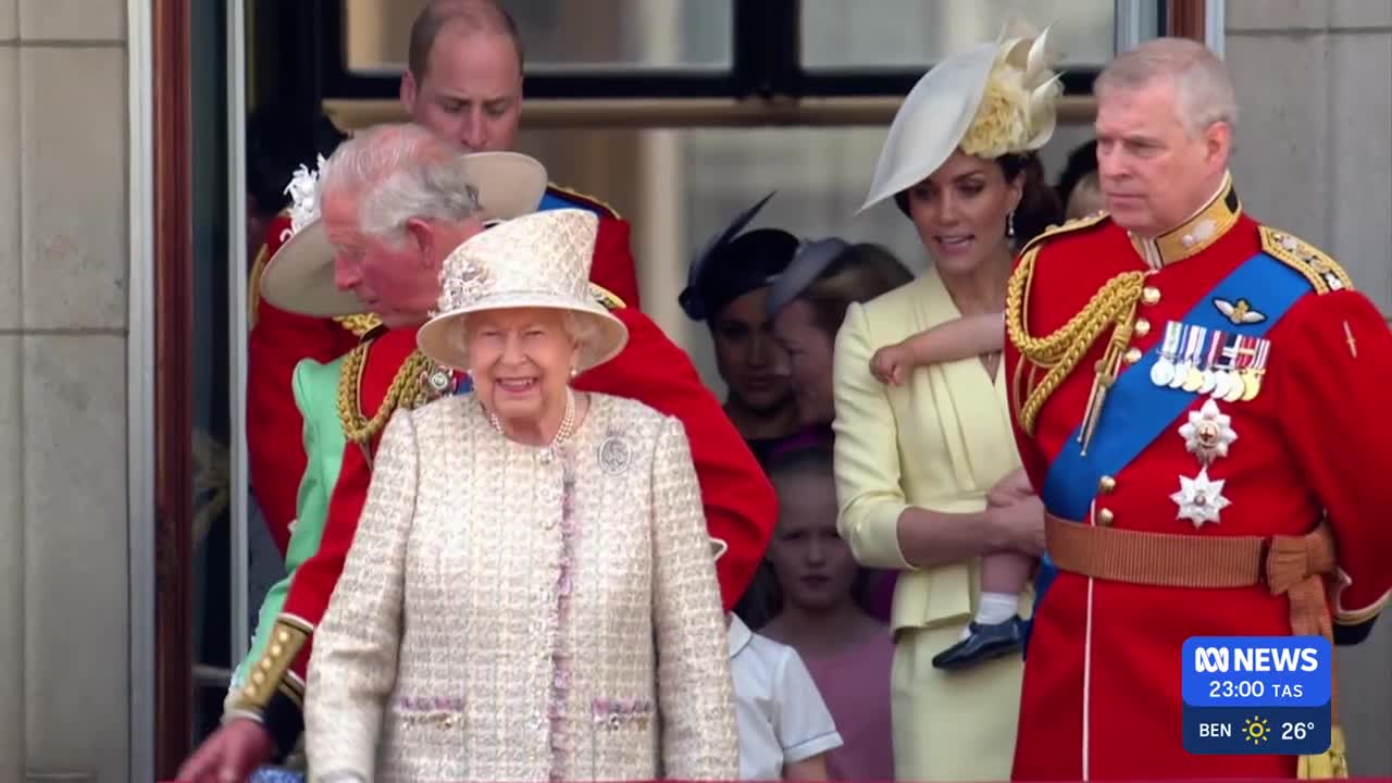 The Queen, in a cream tweed suit and hat, smiles broadly from a balcony. Beside her, Prince Andrew in his red military uniform stands with a stern expression.