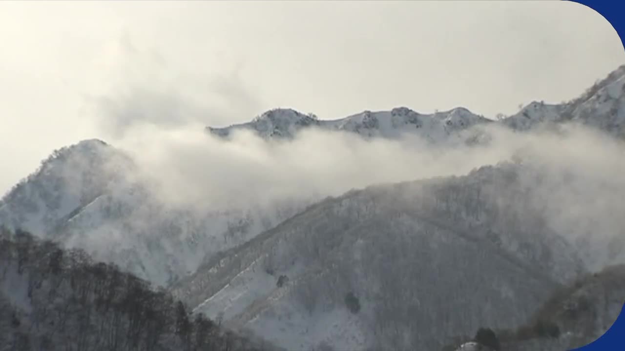 Thick clouds drift through snow-dusted mountain peaks. The trees on the slopes are bare, their branches dusted with white.