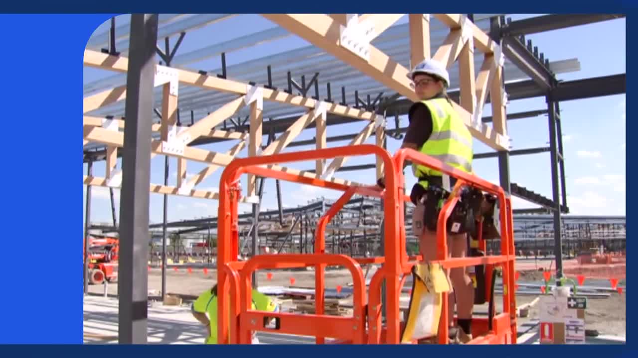 A worker in a bright yellow vest stands in an orange scissor lift, looking out over a construction site. The skeletal frame of a new building rises around them under a clear Australian sky.