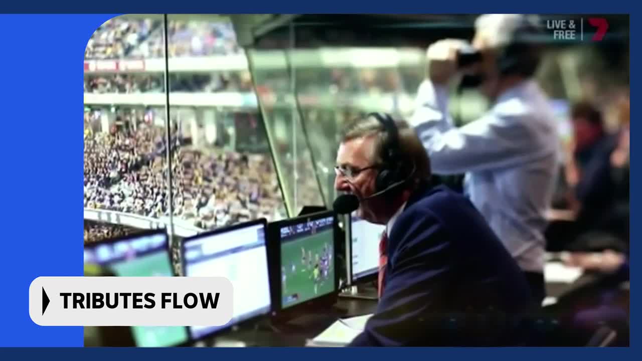 A commentator with a headset leans into his microphone, watching a game unfold on a monitor. The crowd in the stadium behind him is a sea of colour.